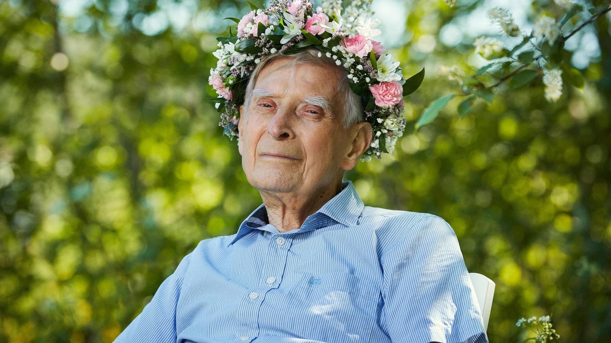 Somrig Blomstedt i randig skjorta med blomsterkrans i håret. Sitter utomhus mot bakgrund av gröna växter.