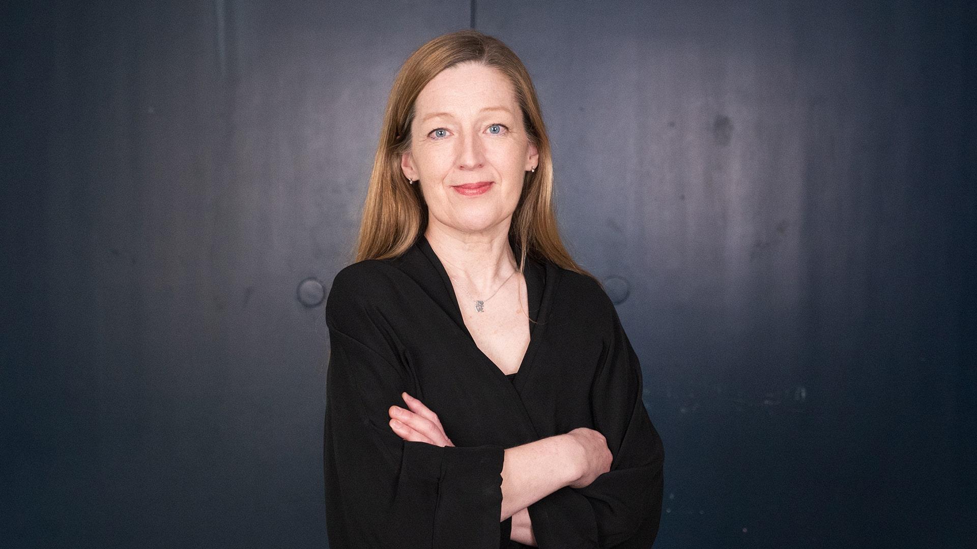 Tove Nilsson, Radiokören. Kvinna med armarna korsade över bröstet tittar rakt in i kameran och ler. Hon har långt hår och är klädd i svart blus och ett silverhalsband.