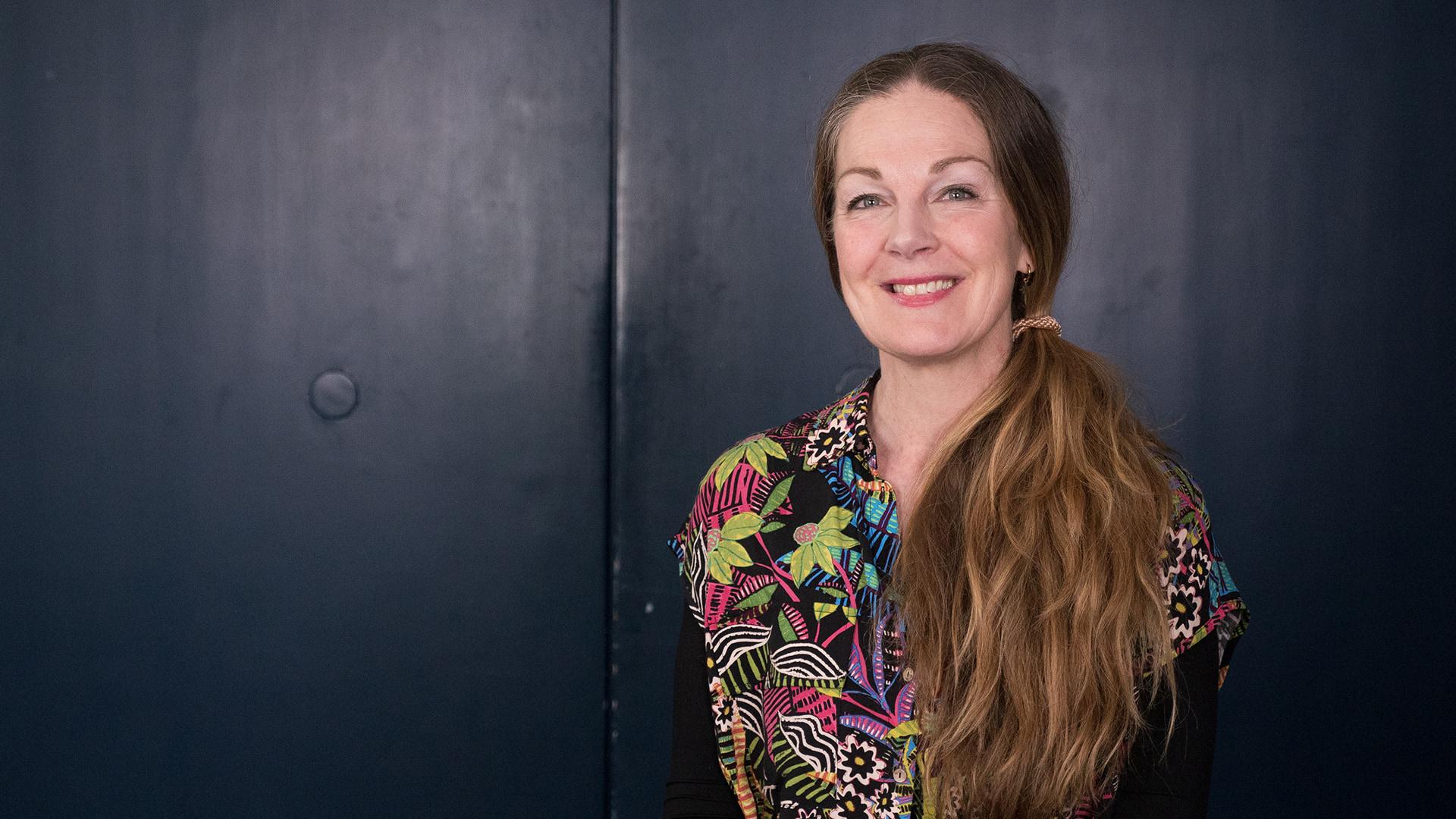 Anna Zander, Radiokören. Leende kvinna med blont långt hår lagt över ena axeln. Hon tittar rakt in i kameran och är klädd i en stormönstrad och färgglad skjorta med blommor och blad.
