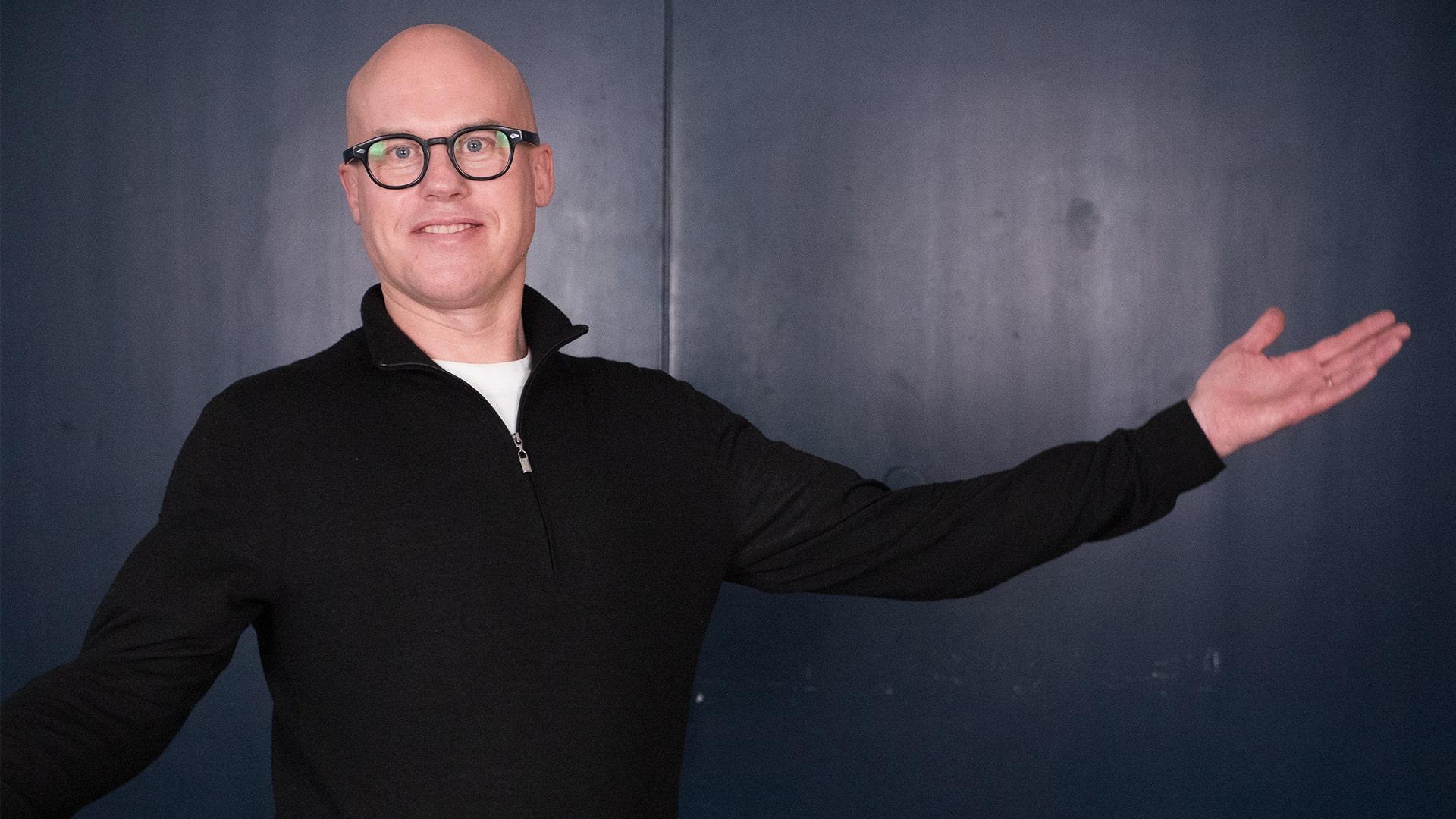 Lars Johansson Brissman, Radiokören. Bild på leende man som sträcker ut armen i en välkomnande gest. Han har svarta glasögonbågar och svart tröja med dragkedja.