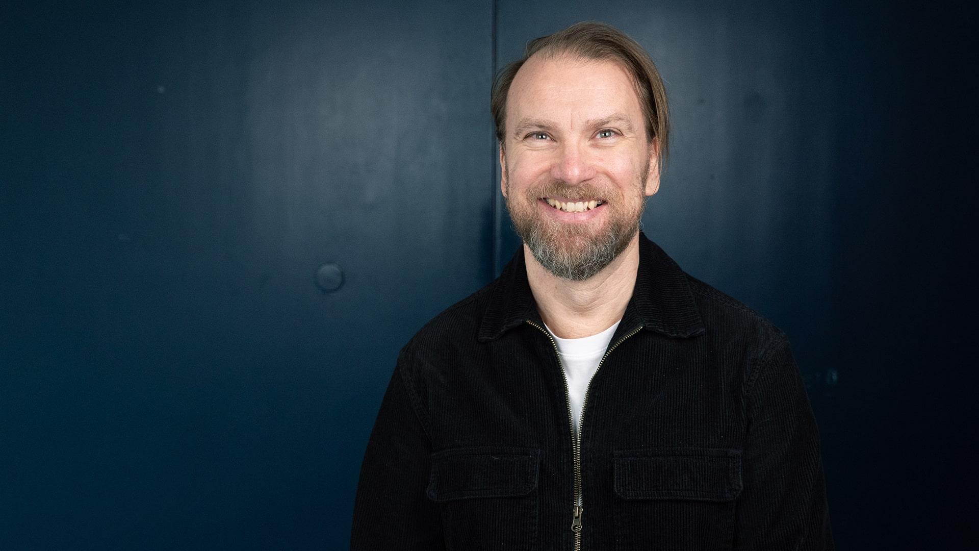 Fredrik Mattson, Radiokören. Leende man tittar rakt in i kameran. Han har skägg och är klädd i svart stickad tröja med vit t-shirt under.