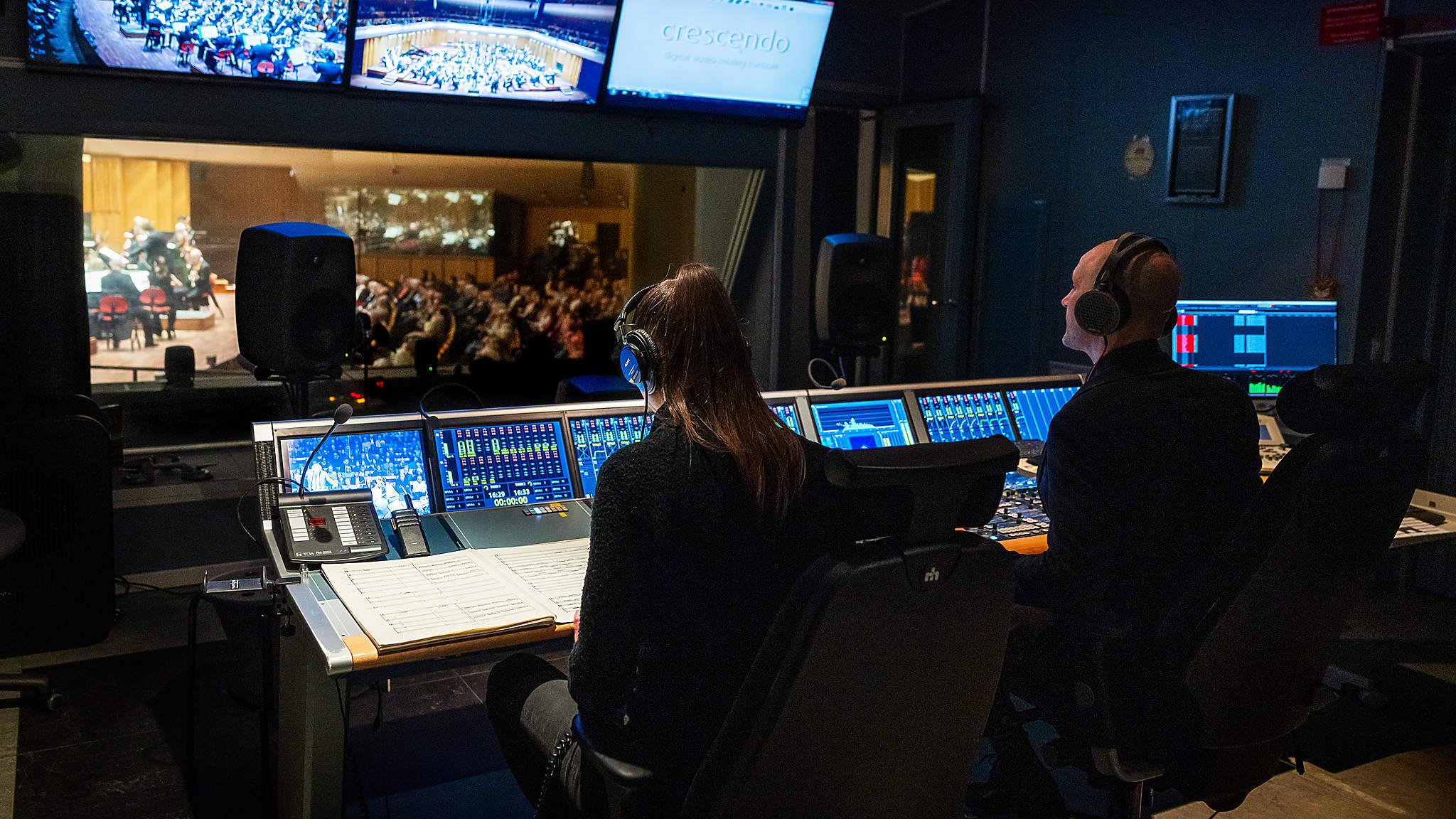 Bild från sändningsrummet i Berwaldhallen. Ljudteknik och skärmar lyser upp rummet där två personer med hörlurar arbetar. I bakgrunden ser man scenen och publiken.