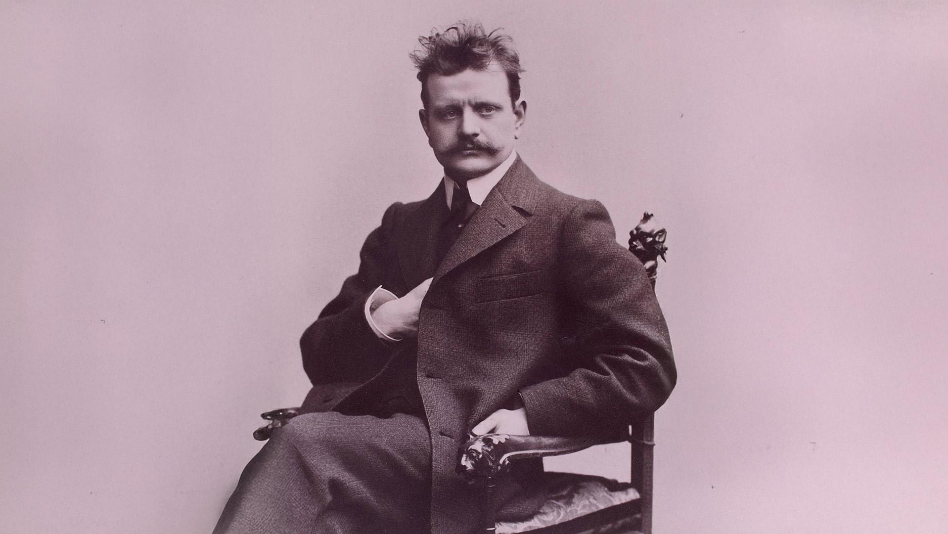 Porträttbild av Jean Sibelius som sitter i en stol med ena handen innanför kavajslaget. Yvigt hår och mustacher.