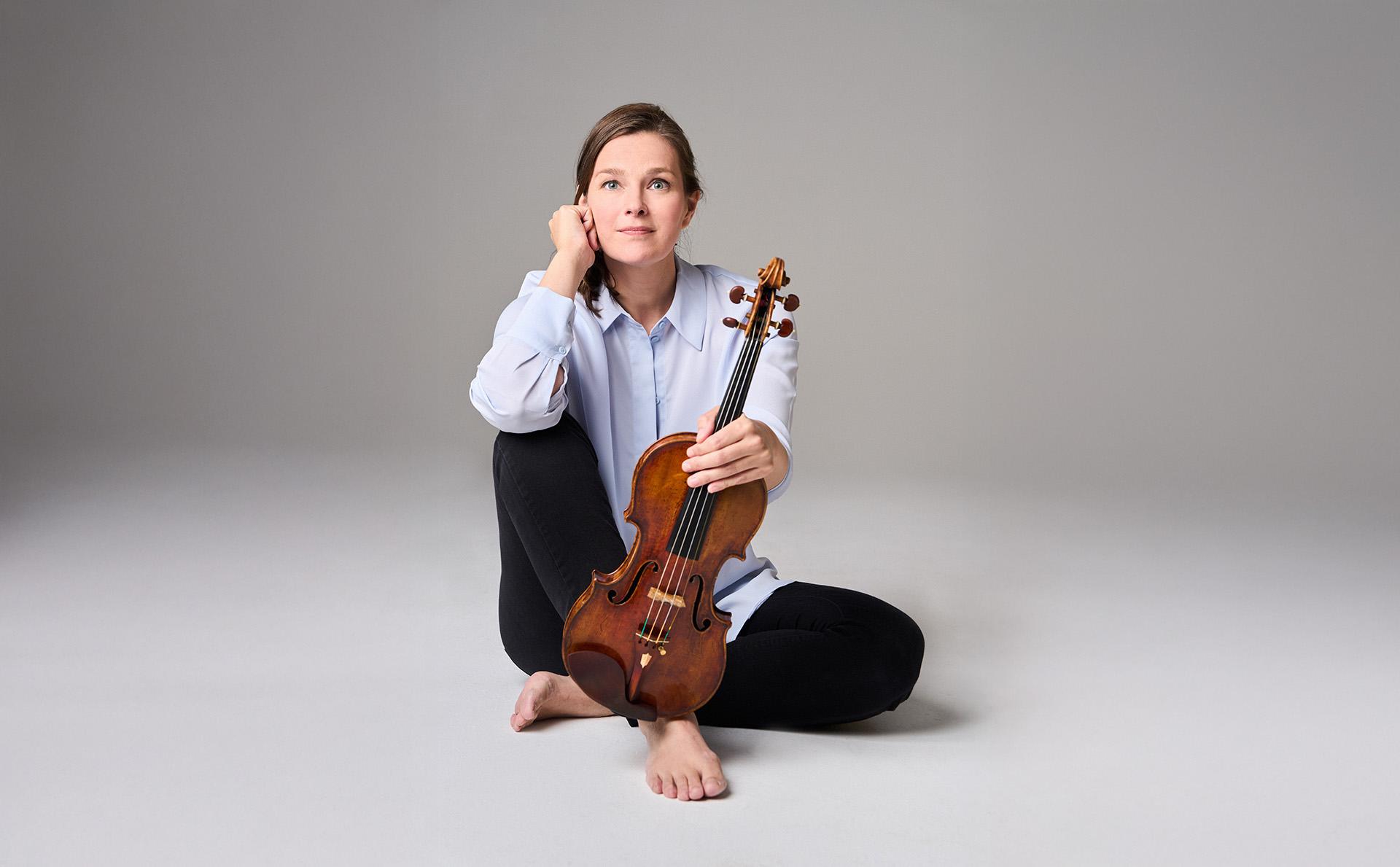Janine Jansen sitter på ett golv och håller sin violin i vänster hand.