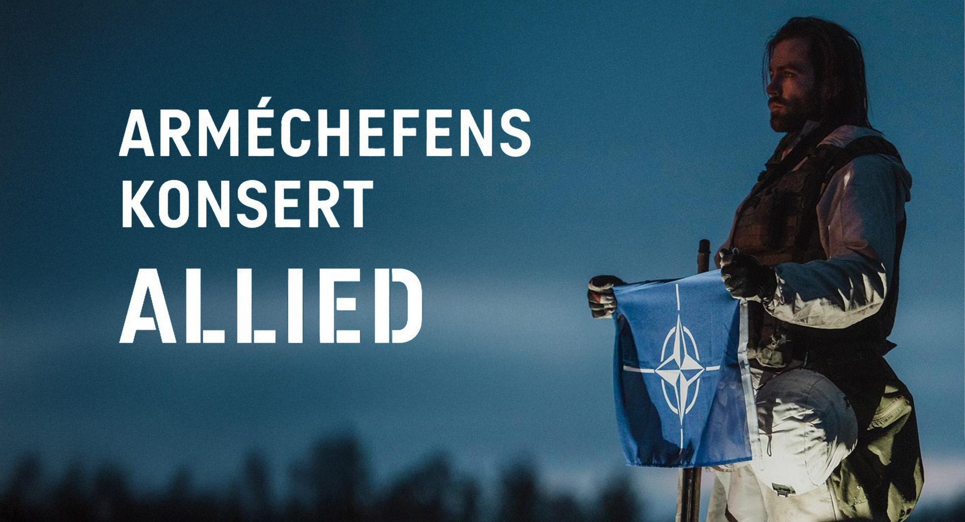 En allvarlig man med axellångt hår i militäruniform blickar ut mot en mörkblå kväll. I händerna håller han NATOs blå flagga med vit kompassrosemblem.